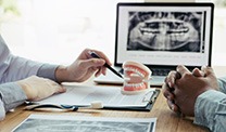 Hands of dentist and patient at desk discussing treatment options next to model teeth and X-rays