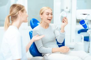 Woman with blond hair in dentist's chair smiling at her reflection in a mirror